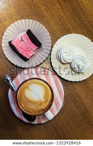 Two white and two pink cakes on two small plates. A cup of cappuccino coffee with a heart on a striped saucer with a spoon. On a wooden table. View from above.