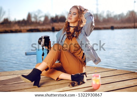 Beautiful girl sitting on the pier near the river. Hipster model in choker with blond cerly hair. The girl at hot sunny day taking pictures old camera, Summer vibes. Woman photographer hobby.