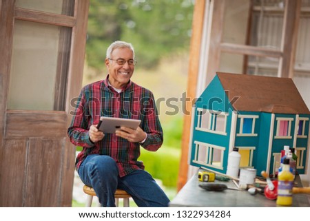 Smiling senior man holding an iPad while sitting in a garage next to a dolls house.