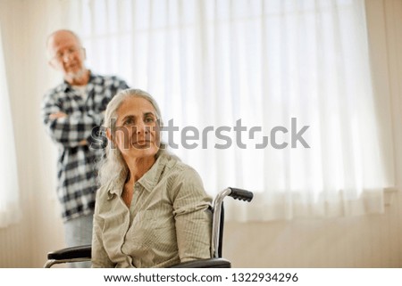 Pensive senior woman sitting in a wheel chair while her husband looks on.