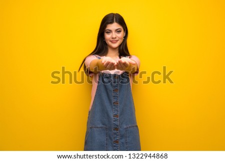 Teenager girl over yellow wall holding copyspace imaginary on the palm to insert an ad