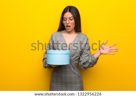 Young woman with glasses over yellow wall holding gift box in hands