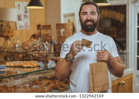 Happy mature bearded man holding shopping bag and credit card, smiling joyfully at the bakery. Cheerful male customer paying for pastry at coffee shop, shopping for food, copy space