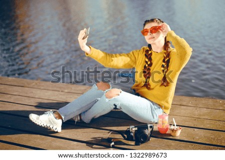 Beautiful girl sitting on the pier near the river. Hipster model yellow with brunette long hair. The girl at hot sunny day taking pictures on phone camera, Summer vibes. Girl take a selfie. Drink