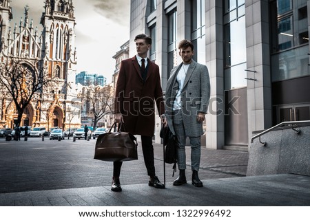 Two fashion men models posing on city street. Wearing in classic and casual closes. Suit, coat, shirt, sweater, boots and leather bag. Men model test near business center Royalty-Free Stock Photo #1322996492