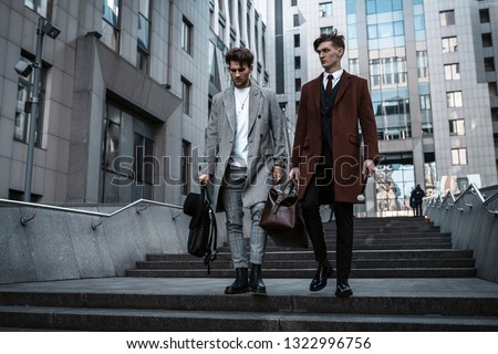 Two fashion men models posing on city street. Wearing in classic and casual closes. Suit, coat, shirt, sweater, boots and leather bag. Men model test near business center Royalty-Free Stock Photo #1322996756