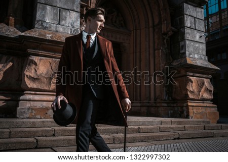 Fashion portrait of young man on red coat, white shirt, black suit and cane walking on streets of city background. Model shooting Royalty-Free Stock Photo #1322997302