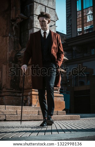 Fashion portrait of young man on red coat, white shirt, black suit and cane walking on streets of city background. Model shooting Royalty-Free Stock Photo #1322998136