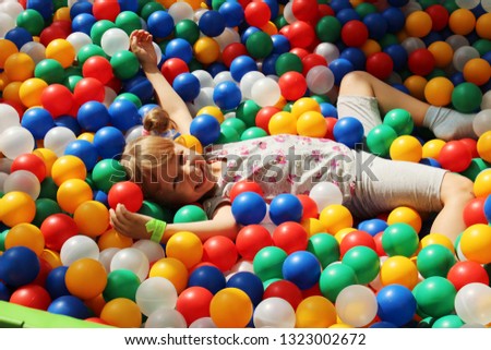 A smiling little girl is lying in a ball pool among colorful plastic balls in the playground