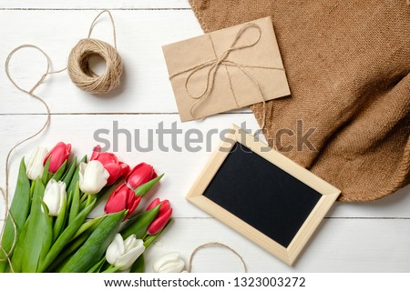 Bouquet of tulips flowers, blank picture frame, kraft envelope, twine, burlap on white wooden table. Vintage greeting card for womans day, mothers day, birthday, easter. Rustic background, flat lay