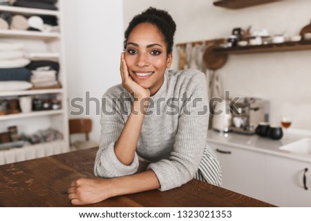Young attractive smiling african american woman with dark curly hair leaning on table happily looking in camera spending time in beautiful cozy kitchen at home   Royalty-Free Stock Photo #1323021353