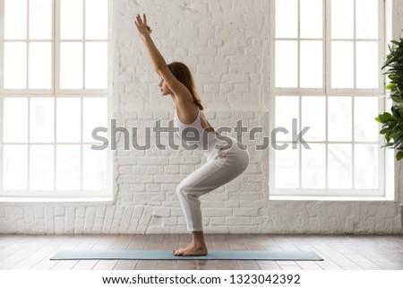 Concentrated woman in white sportswear, pants and top practicing yoga, doing Chair exercise, sporty girl standing in Utkatasana pose in modern yoga studio or at home, side view