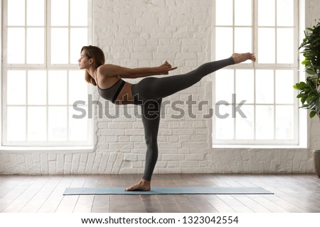 Attractive young woman in grey sportswear, leggings and bra practicing yoga, beautiful girl standing in Warrior three, Virabhadrasana pose, working out at home or in yoga studio with white walls Royalty-Free Stock Photo #1323042554