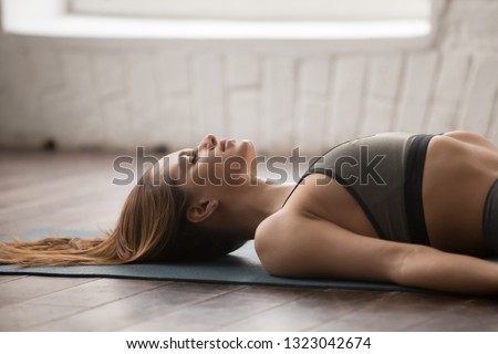 Woman with closed eyes practicing yoga, lying in Savasana, Dead Body pose on mat, beautiful girl in grey sportswear resting after working out at home or in yoga center with white walls, close up Royalty-Free Stock Photo #1323042674