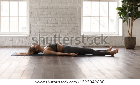 Calm woman in grey sportswear, pants and bra practicing yoga, lying in Savasana, Dead Body pose on mat, beautiful girl resting after working out at home or in yoga center with white walls Royalty-Free Stock Photo #1323042680