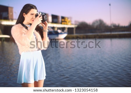 Beautiful girl on the pier near the river. Model in blue dress with long hair. Girl at hot sunny day taking pictures on old camera, Summer vibes. Woman photographer hobby. On the background of yachts