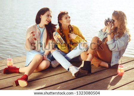 The blonde girl takes pictures of her brunette girlfriends on the old camera. They send her kisses, smile, laugh. Girl with drinks and sunglasses have fun. Girls on the dock/pier on the back of yachts