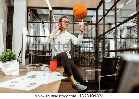 Office games. Joyful nice man throwing the basketball ball while talking on the phone in the office