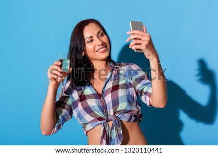 Young woman standing bare belly isolated on blue wall holding glass of champagne taking selfie photos on smartphone smiling joyful