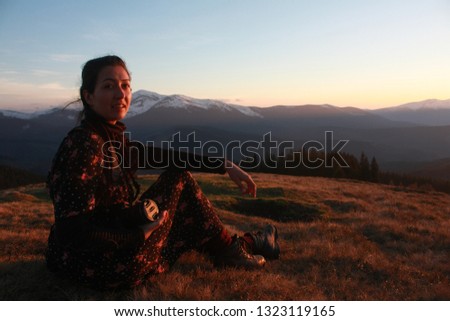 Young beautiful girl in charming dress with long dark hair sitting on a mountain at sunset. Spring mountains, warm colors and twilight
