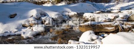 Snow-covered stones and banks with ice edge in the turbulent flow of the winter river on a sunny  day.
