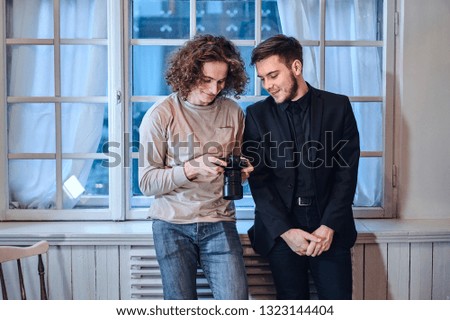 Professional curly haired photographer man showing photos on digital camera to suit dressed and smiling customer at photo studio indoors