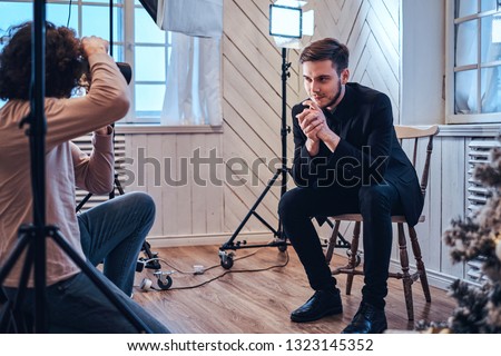 Professional photo shooting at the studio: a handsome young guy sitting, smiling and posing the curly photographer is taking pictures with a digital camera 