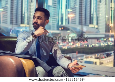Handsome businessman sitting in front of modern urban area full of buildings, holding mobile phone in hand and looking away.