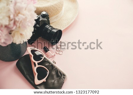 Hello summer.  International womens day. Stylish photo camera, sunglasses, jewelry,straw hat, cosmetics,bag pink and white peonies bouquet on pink paper with space for text.
