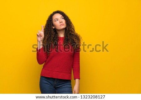 Teenager girl with red sweater over yellow wall with fingers crossing and wishing the best