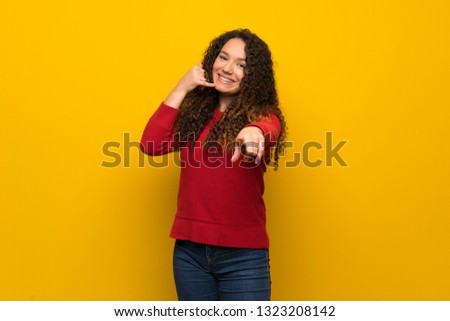 Teenager girl with red sweater over yellow wall making phone gesture and pointing front