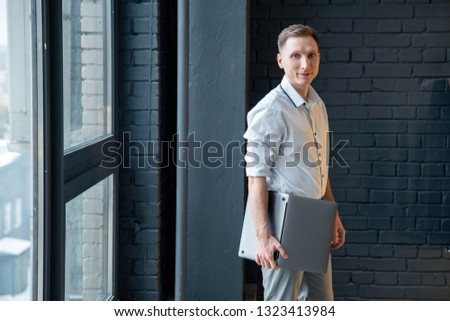 a young man in a white shirt with rolled up sleeves stands at the window and holds a laptop.
