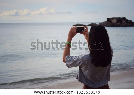 Hippie tourist taking ocean view photo with mobile phone camera on vintage style effect, free space. Hipster girl shooting vdo of sea beach with white clouds sky at tropical island, blurred background