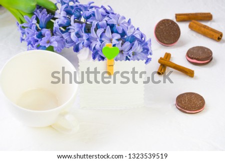 White empty mug on a background of spring flowers. Blue hyacinth lies in the background, a number of dishes and chocolate chip cookies, still life adorn cinnamon sticks. Nearby there is a paper card f