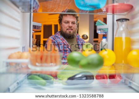 Man looking for something to eat at night while standing in front of opened fridge. Unhealthy eating concept. Picture taken from the inside of fridge.