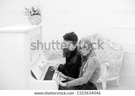 Young couple in enormous glasses sitting in white room and playing piano. Stylish beaded man and his blond girlfriend crazy sitting at grand piano.