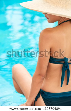 Rear view of beautiful woman in hat sitting near the swimming pool and sunbathing outdoors at resort