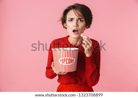 Portrait of a confused beautiful young woman wearing red dress standing isolated over pink background, eating popcorn