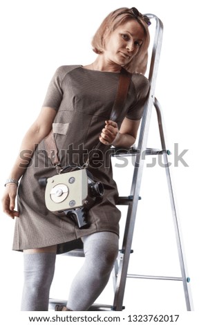 Young woman in dress and with retro movie camera posing on stepladder isolated on white background.