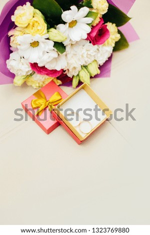 
Two gold wedding rings are in a small square box with a banton on a beige background next to the bride's bouquet of flowers