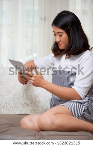 Asian young girl in dress sitting on sofa and working online using digital tablet at home