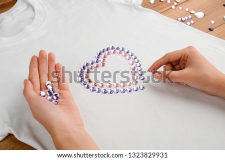 Girl laying out a heart of violet and pink rhinestones on a white t-shirt, close up. Simple and beautiful custom design for clothes. Royalty-Free Stock Photo #1323829931