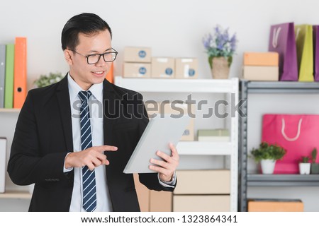 Close up portrait of young handsome Asian businessman wearing eyeglasses in dark suit, working in modern startup office using tablet browsing data on marketing information