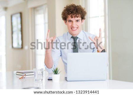 Young business man working with computer laptop at the office shouting with crazy expression doing rock symbol with hands up. Music star. Heavy concept.