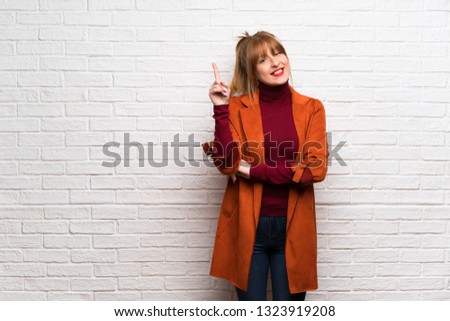 Woman with coat over white brick wall showing and lifting a finger in sign of the best