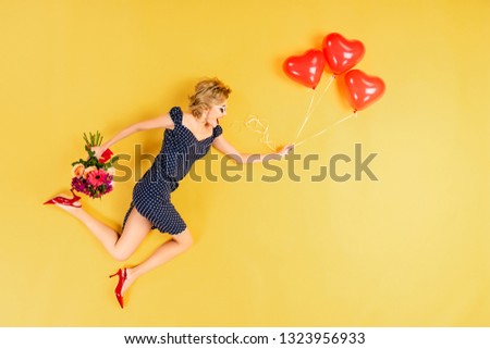 Excited woman with heart-shaped air balloons and flowers jumping on yellow background