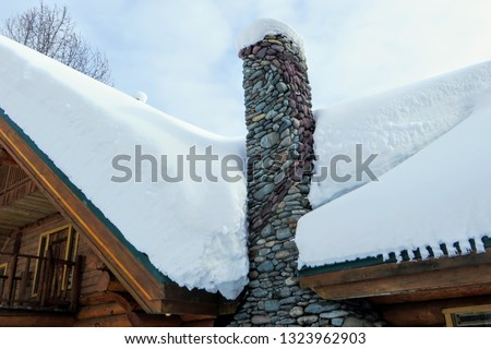 A closeup view of a stone chimney attached to a remote log cabin covered in freshly fallen snow.