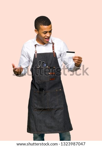 Young afro american barber man holding a credit card and surprised on isolated background