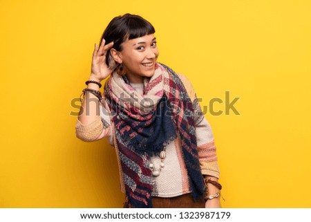 Young hippie woman over yellow wall listening to something by putting hand on the ear