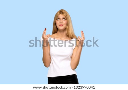 Young blonde woman with fingers crossing and wishing the best on isolated blue background